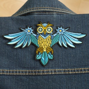 May include: An embroidered owl patch in blue and yellow with intricate details. The owl has large, round eyes and outstretched wings. The patch is sewn onto a denim jacket.