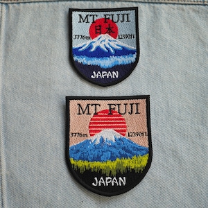 Könnte beinhalten: Zwei gestickte Patches mit dem Motiv des Mount Fuji, Japan. Beide Patches haben die Form eines Schildes und zeigen eine rote Sonne, die über dem Berg aufgeht. Der obere Patch hat einen blauen Hintergrund und der untere Patch einen rosa Hintergrund. Beide Patches zeigen den Text "MT FUJI" und "JAPAN" in Schwarz.