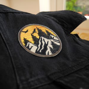 May include: Circular embroidered patch depicting a mountain scene with trees, birds, and a sunset. The patch, with a brown border, is sewn onto a black denim jacket. The design incorporates yellow, orange, black, and white.