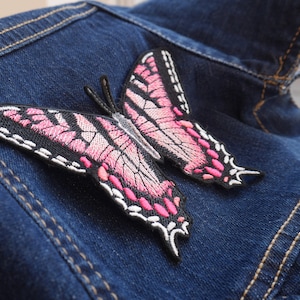 Könnte beinhalten: Rosa-schwarzer gestickter Schmetterlings-Patch auf einer blauen Jeansjacke.