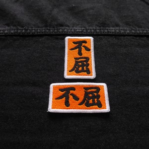 K&ouml;nnte beinhalten: Zwei orangefarbene Stoffpatches, die auf eine schwarze Jeansjacke gen&auml;ht sind. Die Patches haben wei&szlig;e R&auml;nder und schwarzen Text, der in chinesischen Schriftzeichen "不屈" lautet.