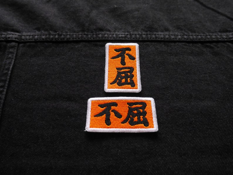 K&ouml;nnte beinhalten: Zwei orangefarbene Stoffpatches, die auf eine schwarze Jeansjacke gen&auml;ht sind. Die Patches haben wei&szlig;e R&auml;nder und schwarzen Text, der in chinesischen Schriftzeichen "不屈" lautet.