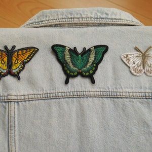 May include: Three butterfly patches in yellow, green, and white, with detailed wing patterns, are displayed on a light blue denim jacket. These embroidered patches offer a way to personalise clothing and add a touch of nature.