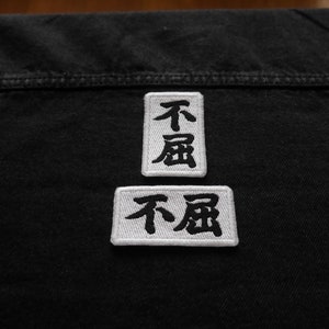 K&ouml;nnte beinhalten: Zwei wei&szlig;e Stoffpatches mit schwarzen gestickten japanischen Schriftzeichen, "不屈", was "unbeugsam" oder "unbezwingbar" bedeutet, auf eine schwarze Jeansjacke gen&auml;ht.