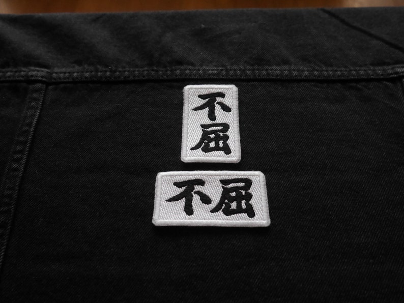 K&ouml;nnte beinhalten: Zwei wei&szlig;e Stoffpatches mit schwarzen gestickten japanischen Schriftzeichen, "不屈", was "unbeugsam" oder "unbezwingbar" bedeutet, auf eine schwarze Jeansjacke gen&auml;ht.
