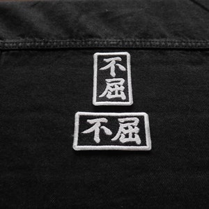 K&ouml;nnte beinhalten: Zwei schwarze und wei&szlig;e gestickte Patches auf einer schwarzen Jeansjacke. Die Patches sind rechteckig und zeigen die chinesischen Schriftzeichen f&uuml;r "nicht" und "Sorge".