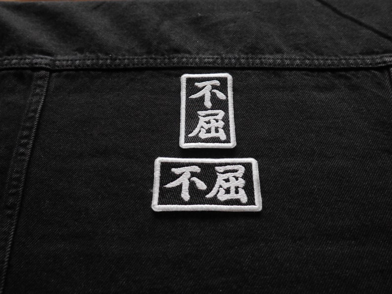 K&ouml;nnte beinhalten: Zwei schwarze und wei&szlig;e gestickte Patches auf einer schwarzen Jeansjacke. Die Patches sind rechteckig und zeigen die chinesischen Schriftzeichen f&uuml;r "nicht" und "Sorge".