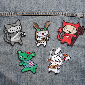 May include: Five embroidered iron-on patches featuring cartoon animals. The patches are on a blue denim background. The patches include a black and white cat wearing a ninja outfit, a white rabbit wearing goggles and a pilot's hat, a red devil with a pitchfork, a green zombie bear, and a white rabbit wearing an eye patch and a pirate hat.