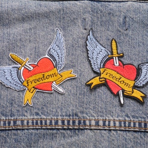 Op de afbeelding: Twee geborduurde patches met een rood hart dat door een zwaard met vleugels wordt doorboord en een banner met de tekst "Freedom".