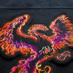 Può includere: Una giacca di jeans nera con una toppa ricamata a forma di fenice. La fenice è raffigurata in tonalità di arancione, rosso e viola, con dettagli intricati nelle sue ali e nella sua coda.