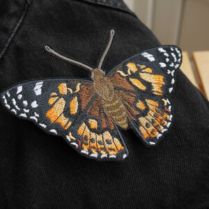 May include: Embroidered butterfly patch with detailed wings in shades of orange, brown, black, and white. The patch is attached to a black denim jacket. The butterfly has a detailed body and antennae.