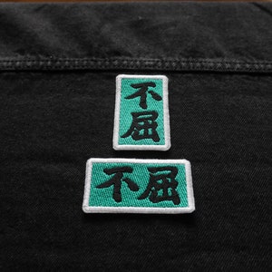 K&ouml;nnte beinhalten: Zwei gestickte Patches in Schwarzwei&szlig; auf einem schwarzen Jeansstoff. Die Patches sind rechteckig und haben einen t&uuml;rkisfarbenen Rand. Der Text auf den Patches lautet "不屈".
