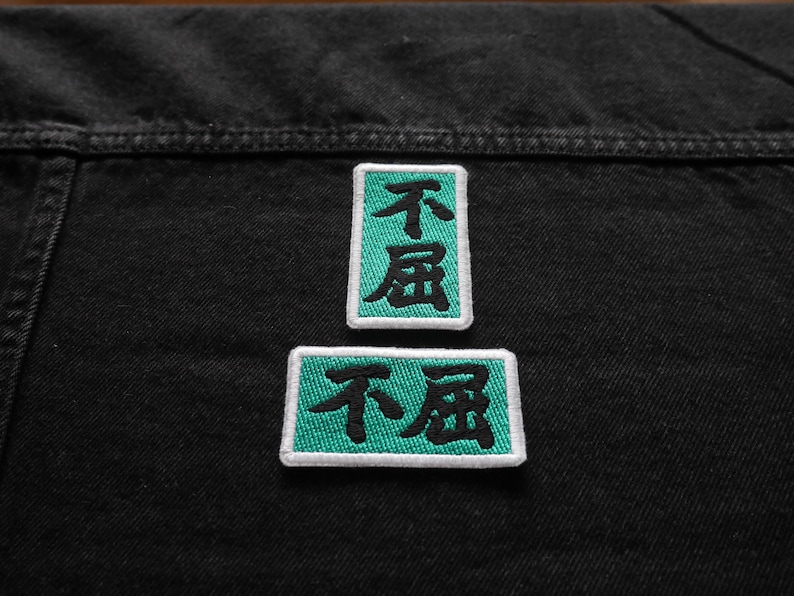 K&ouml;nnte beinhalten: Zwei gestickte Patches in Schwarzwei&szlig; auf einem schwarzen Jeansstoff. Die Patches sind rechteckig und haben einen t&uuml;rkisfarbenen Rand. Der Text auf den Patches lautet "不屈".