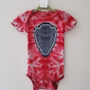 Op de afbeelding: Rode, witte en grijze tie-dye baby romper met een grijze pijl in het midden. De romper heeft een label met de tekst "12m".