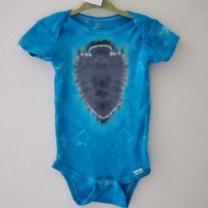 Op de afbeelding: Een blauwe tie-dye baby onesie met een grijs hartvormig ontwerp. De onesie heeft een label met de tekst "12m".