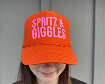 May include: Orange trucker hat with pink text that reads "SPRITZ & GIGGLES".