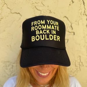 Puede incluir: Una gorra de camionero negra con una malla amarilla en la parte posterior. La gorra tiene el texto "FROM YOUR ROOMMATE BACK IN BOULDER" impreso en amarillo en la parte delantera.