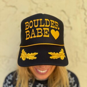 Puede incluir: Gorra de camionero negra con bordado dorado. La gorra dice "BOULDER BABE" con un corazón dorado.