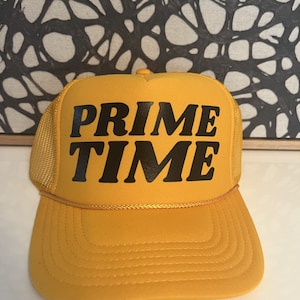Puede incluir: Gorra de camionero de color amarillo mostaza con las palabras "PRIME TIME" en letras negras en negrita en la parte delantera. La gorra tiene una parte trasera de malla y una visera curva. Un detalle de cuerda dorada acentúa la visera.