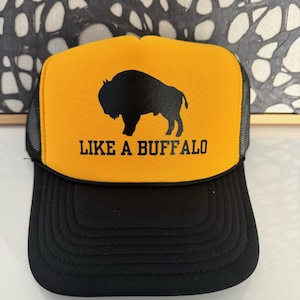Puede incluir: Una gorra de camionero amarilla y negra con una silueta negra de búfalo y el texto "LIKE A BUFFALO" en negro.