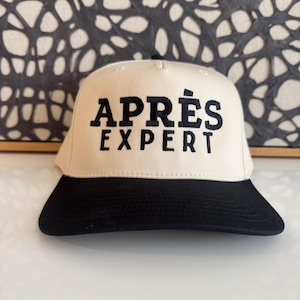 May include: A baseball cap with a cream-colored crown and a black brim. The front of the crown features the text "APRÈS EXPERT" embroidered in bold, black, capital letters. The cap has a structured front panel and a flat, stitched brim design.