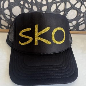 May include: Black trucker hat with the word "SKO" in gold glitter lettering across the front. The hat has a mesh back and a curved brim. The hat is a classic style.