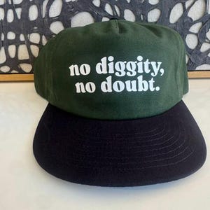 May include: A forest green baseball cap with a flat black brim. The front of the cap features the white text "no diggity, no doubt." printed in a bold, rounded font. The cap has a structured crown and a classic five-panel design.