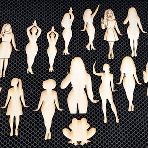 Könnte beinhalten: Holzausschnitte verschiedener weiblicher Figuren in unterschiedlichen Posen, zusammen mit Blatt- und Froschformen. Die Ausschnitte sind aus hellem Holz gefertigt und auf einer dunklen, strukturierten Oberfläche angeordnet.