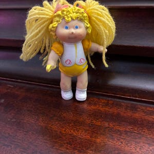 May include: A vintage Cabbage Patch Kids doll with blonde yarn pigtails and blue eyes. The doll wears a yellow and white outfit with a floral design and white shoes. The doll is a collectible toy.