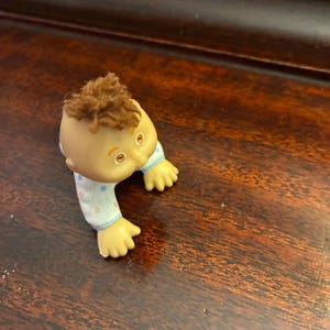 Op de afbeelding: Een kleine plastic babypop met een gebruind gezicht, bruin haar en blauwe ogen. De pop draagt een lichtblauwe en witte polkadot onesie. De pop kruipt.