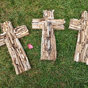 May include: Three wooden crosses crafted from driftwood, arranged in a row on a bed of green grass. The crosses are all the same size and shape, and they are made from a variety of different pieces of driftwood.