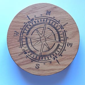 May include: A circular wooden coaster featuring a compass design etched into the surface. The compass rose is central, with directional markers for North, South, East, and West. The wood grain is visible, giving it a natural appearance.