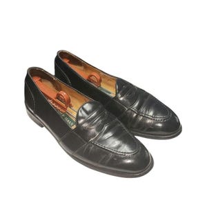 May include: A pair of black leather penny loafers with a moccasin-style stitched toe, a low heel, and a strap across the vamp. The interior features a tan lining and a wooden shoe tree with a metal handle, with the brand name Cole Haan visible.