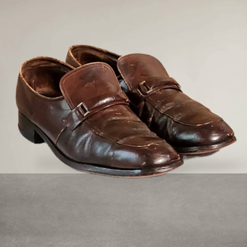 Puede incluir: Un par de zapatos marrones de cuero con un detalle de hebilla. Los zapatos tienen una punta ligeramente puntiaguda y un tac&oacute;n bajo.