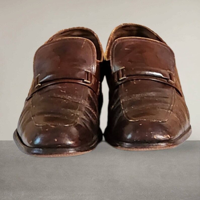 Puede incluir: Un par de zapatos de vestir de cuero marr&oacute;n con un detalle de hebilla. Los zapatos tienen una parte superior de cuero texturizado y un acabado pulido.