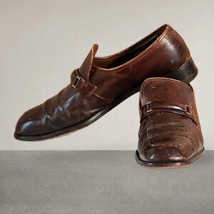 Puede incluir: Un par de zapatos marrones de cuero con un detalle de hebilla. Los zapatos tienen un aspecto ligeramente desgastado y est&aacute;n hechos de cuero texturizado.