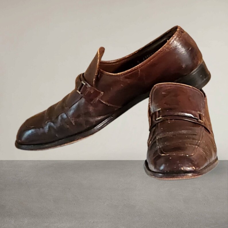 Puede incluir: Un par de zapatos marrones de cuero con un detalle de hebilla. Los zapatos tienen un aspecto ligeramente desgastado y est&aacute;n hechos de cuero texturizado.