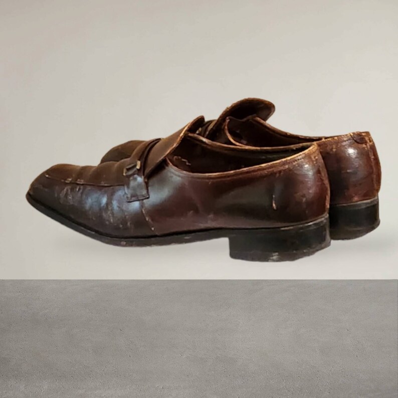 Puede incluir: Un par de mocasines de cuero marr&oacute;n con un detalle de hebilla. Los zapatos tienen un aspecto desgastado y est&aacute;n ligeramente desgastados.