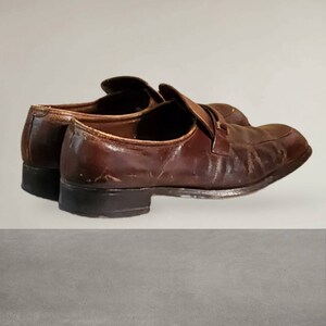 Puede incluir: Un par de mocasines de cuero marr&oacute;n con una correa en la parte superior. Los zapatos tienen un aspecto desgastado y est&aacute;n ligeramente desgastados.