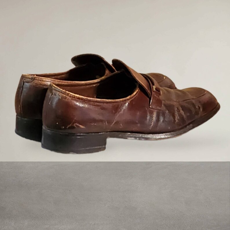 Puede incluir: Un par de mocasines de cuero marr&oacute;n con una correa en la parte superior. Los zapatos tienen un aspecto desgastado y est&aacute;n ligeramente desgastados.