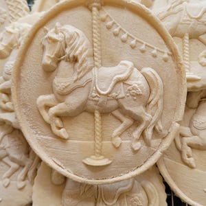 May include: A round, light beige cookie featuring a detailed embossed design of a carousel horse. The horse is depicted in a prancing pose, attached to a central pole with decorative bunting hanging above it, set against a circular border.