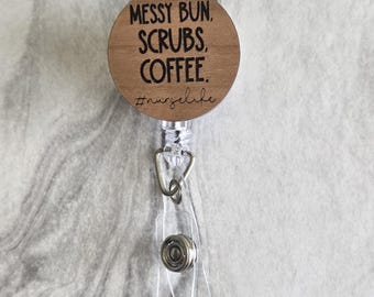 Può includere: Un porta badge in legno con la scritta "MESSY BUN, SCRUBS, COFFEE" e l'hashtag "#nurselife". Il porta badge ha un cordino retrattile in plastica trasparente e una clip in metallo.
