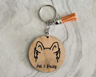 Può includere: Un portachiavi in legno con una silhouette di cane e il testo "Jax & Daisy" inciso su di esso. Il portachiavi ha un anello portachiavi argentato e un piccolo pon pon arancione.