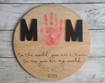 May include: A round wooden sign with the word "MOM" in black letters, a pink handprint, and the text "to the world you are a mom. to me you are my world. LOVE STELLA 2023".
