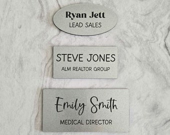 Può includere: Tre targhette porta-nome argentate con testo nero. La targhetta superiore recita "Ryan Jett, Responsabile vendite". La targhetta centrale recita "Steve Jones, Gruppo Immobiliare ALM". La targhetta inferiore recita "Emily Smith, Direttore Medico".