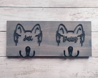 Può includere: Un porta guinzagli per cani in legno con due ganci. Il porta guinzagli è dipinto di un colore marrone scuro e ha due sagome di cani con i nomi "Jax" e "Daisy" ritagliati.
