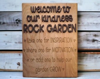 Può includere: Cartello in legno con il testo "Benvenuti nel nostro giardino di rocce della gentilezza" e istruzioni per prendere, condividere o aggiungere una roccia per ispirazione, motivazione o per aiutare il giardino a crescere.