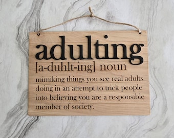 Può includere: Un cartello di legno con la parola "adulting" definita come "imitare le cose che vedi fare agli adulti reali nel tentativo di ingannare le persone facendole credere che sei un membro responsabile della società."