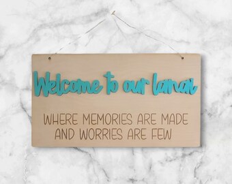Può includere: Un cartello di legno con il testo "Welcome to our lanai where memories are made and worries are few" in lettere blu turchese.