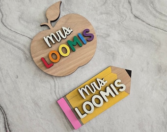 Può includere: Due cartelli di legno con il testo "Mrs. Loomis" su di essi. Un cartello ha la forma di una mela con lettere arcobaleno. L'altro cartello ha la forma di una matita con lettere gialle.